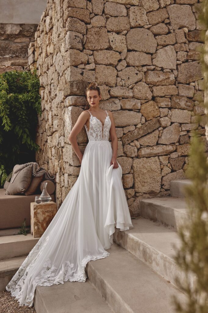 66363-Lillian West-Pure Bridal, Chiffon A-line gown with plunging V-neckline with mesh insert and delicate lace straps. Sheer bodice features Chantilly lace base with exposed boning peeking out. Overlayed with matte lace appliques throughout bodice and onto skirt waistline. Features mesh V side cut-outs and an open V-back with buttons to waist. Chiffon skirt trails into chapel length train with detailed lace cut-outs at train hem. Edmonton wedding shop, Edmonton bridal shop, Edmonton wedding dress.
