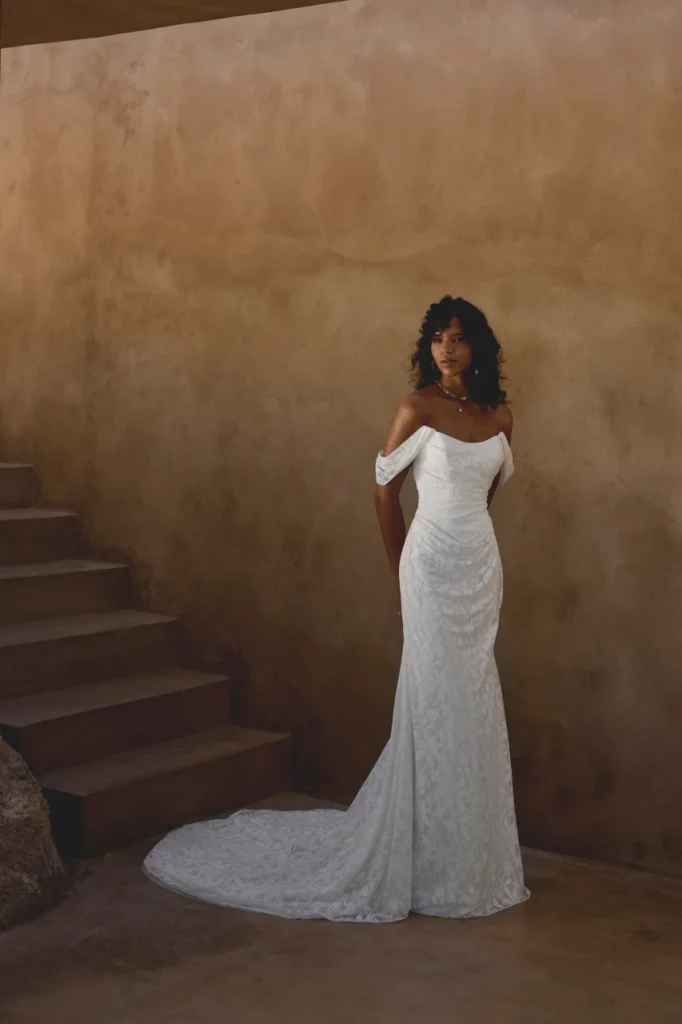 Lillian West style 66442 stretch jacquard wedding dress with an organic floral pattern, featuring a strapless cat eye neckline, ruched bodice, and off-the-shoulder straps, seen in 66442-Lillian West-Pure Bridal.webp. This fit and flare gown with a chapel length train is available at Pure Bridal in Edmonton.
