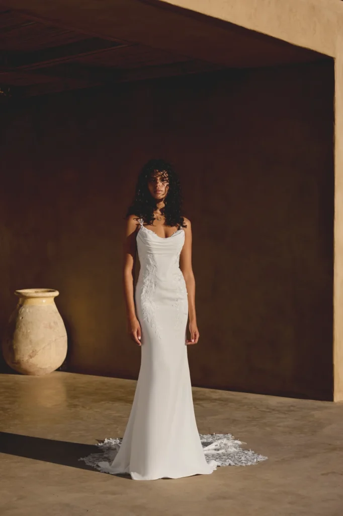 Lillian West style 66447 is a Crepe fitted gown with a bikini neckline featuring thin spaghetti straps. Sequined and beaded lace appliques adorn the bodice with tonal seed beads and pearls and cascade onto the skirt. The front lined bodice also features a draped crepe detail across the bust and a basque waistline and makes way to an illusion back bodice. Crepe covered buttons extend down the chapel length train finished with a detailed cut out train with tulle and lace applique. Available at Pure Bridal Edmonton.