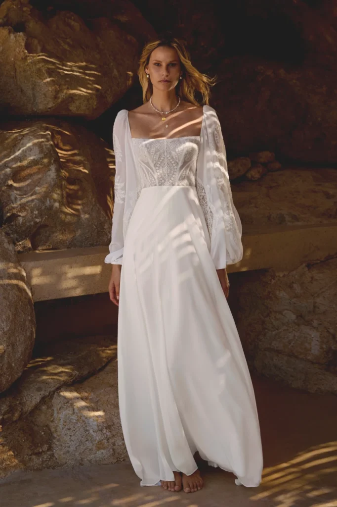 Lillian West style 66461 boho A-line wedding dress in chiffon, featuring a sheer bodice with exposed boning, a square neckline, and long blouson sleeves with matte lace cut-outs. Finished with a chapel length train and available at Pure Bridal in Edmonton.