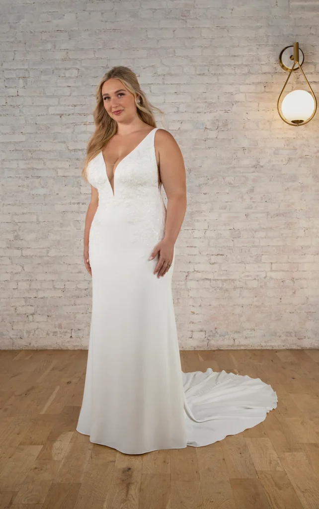 Style 7774 by Stella York, a glamorous luxury crepe column wedding dress with a deep plunging neckline, beaded lace appliqu&eacute;s, modern side cutouts, and a low-scooped back, available in plus sizes at Pure Bridal Edmonton bridal boutique.