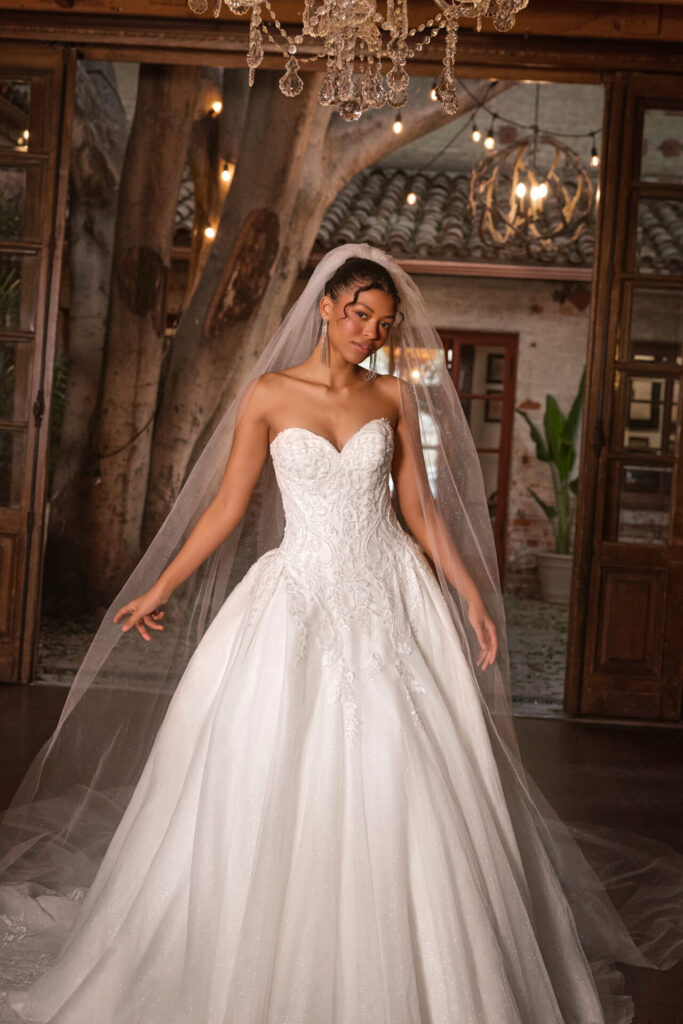D4331-Essense of Australia-Pure Bridal. Classic bridal elegance meets modern glamour in this breathtaking ballgown. Style D4331 features a beautifully embellished sweetheart neckline highlighting the decolletage, adding a touch of romance to the structured bodice. The grand silhouette comes to life with shimmering glitter tulle, while pleated layers create soft movement and dimension. Delicate scrolls of lace cascade throughout, adding a regal yet ethereal touch. For a timeless finish, fabric-covered buttons trace from the top of the neckline down to the edge of the sweeping train. This showstopping gown is also available in plus sizes. Edmonton wedding shop, Edmonton bridal shop, Edmonton wedding dress.