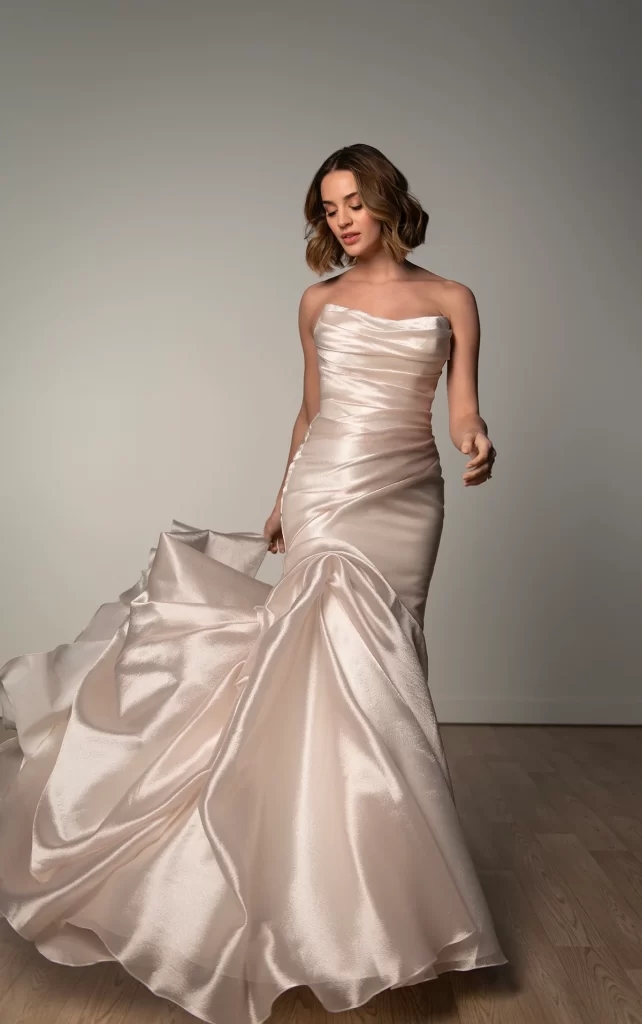 SY8123-Stella York-Pure Bridal. Edmonton wedding dress. Edmonton Bridal shop. Style SY8123 is a striking showcase of modern sculptural design, ideal for the fashion-forward bride. This bold fit-and-flare wedding gown features a sleek strapless bodice with a softly scooped cat-eye neckline and elegant ruching throughout. A trail of fabric-covered buttons cascades down the back, leading to a voluminous asymmetrical skirt with glamorous pickups that add dynamic movement. Effortlessly blending clean lines with dramatic flair, this head-turning design embodies couture craftsmanship, offering a chic and contemporary bridal look that is both sophisticated and unforgettable. This gown is also available in plus sizes.
