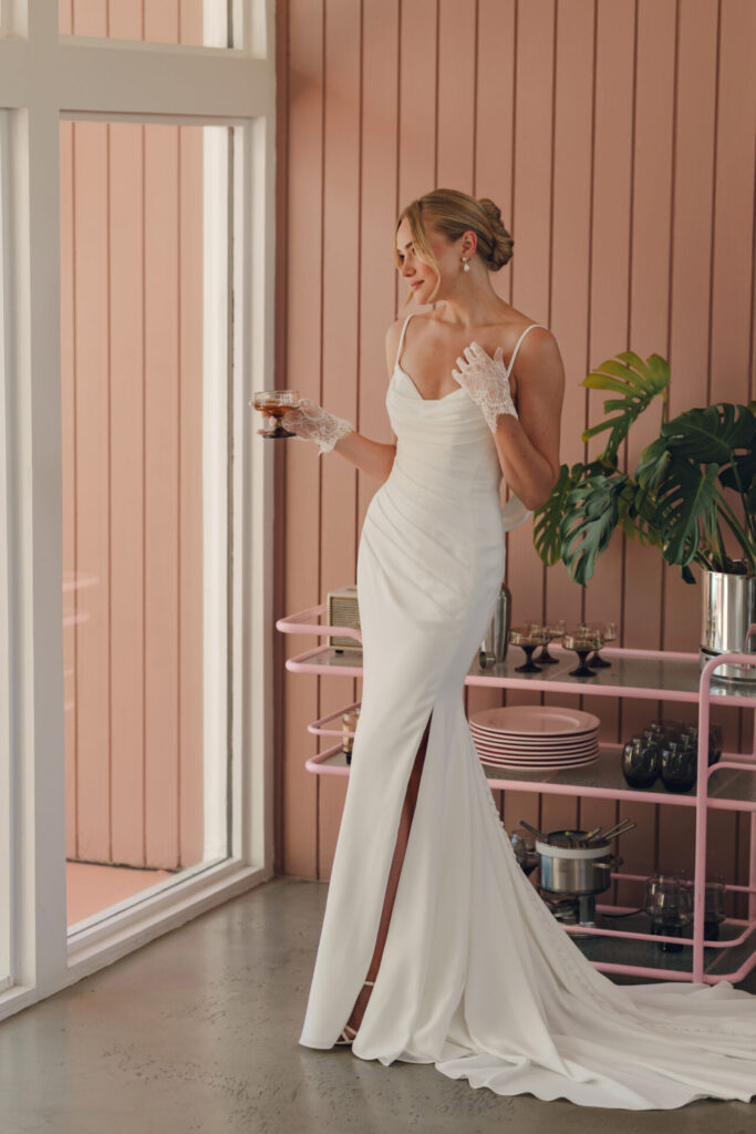 Style SY8224 by Stella York, a chic crepe column wedding dress with a draped scoop neckline, dainty spaghetti straps with a detachable back cowl, asymmetrical ruching, and a high leg slit available in plus sizes at Pure Bridal boutique in Edmonton.
