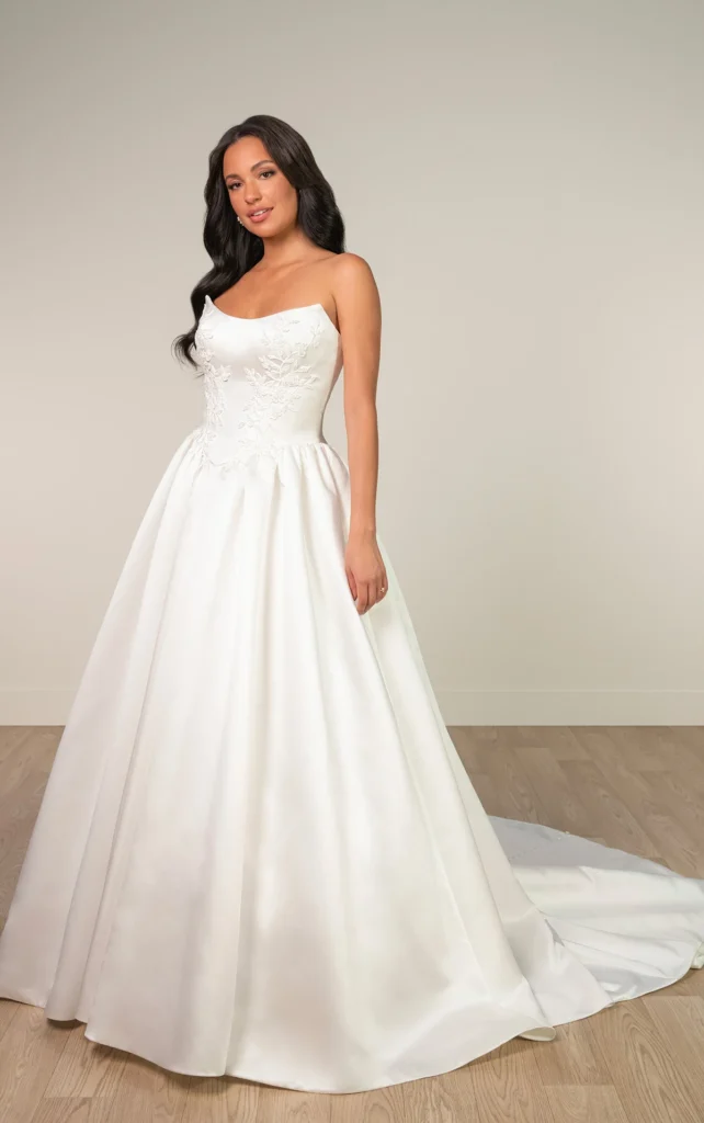 Style SY8239 by Stella York, a classic Grand Satin ballgown wedding dress with a strapless cat-eye neckline, lace-appliqu&eacute;d bodice, and a basque waist, with sculpted pleats and a voluminous skirt, available in plus sizes at Pure Bridal boutique in Edmonton.