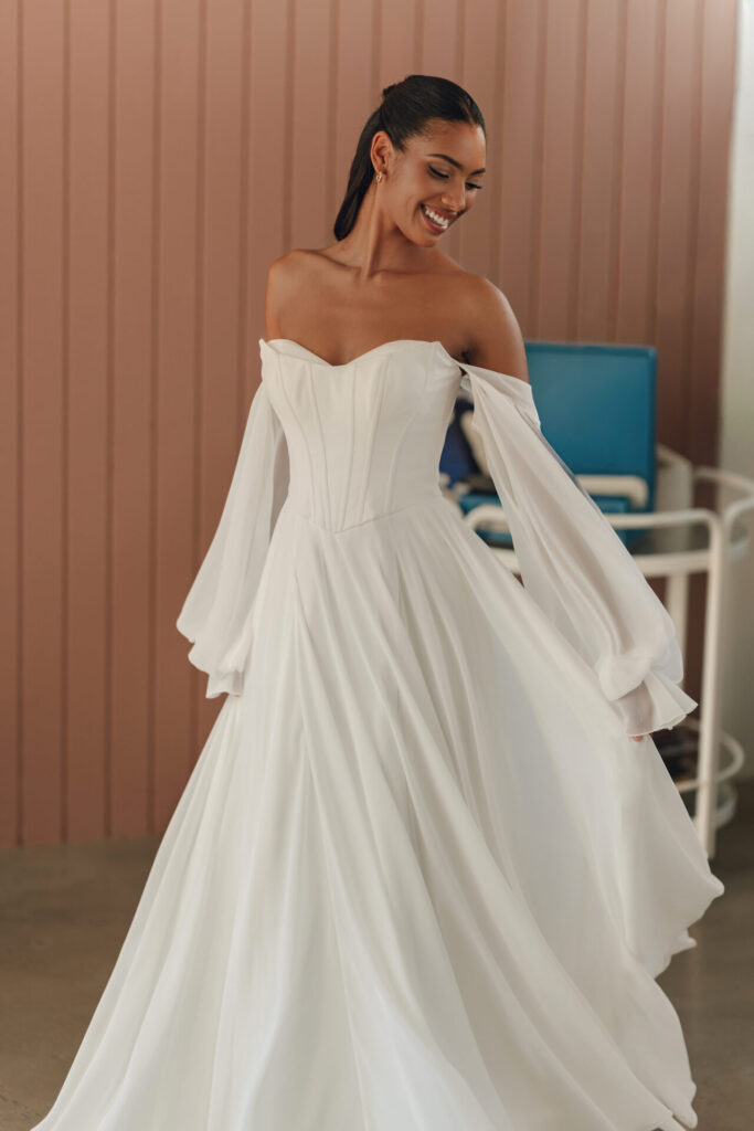 Style SY8244 by Stella York, a romantic chiffon A-line wedding dress with a structured corset bodice, a basque waist, off-the-shoulder long detachable blouson sleeves, with a flowing skirt and a high leg slit, available in plus sizes at Pure Bridal boutique in Edmonton.