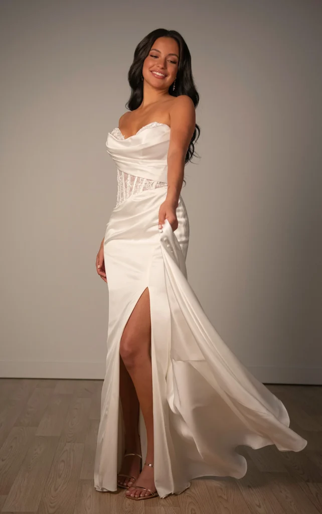 Style SY8248 by Stella York, a modern Belle Satin column wedding dress with a strapless draped bodice, an asymmetrical peekaboo structured lace-appliqued corset, and a high leg slit, available at Pure Bridal boutique in Edmonton.