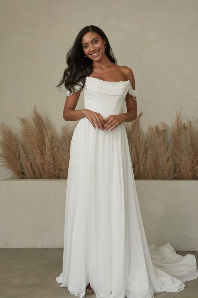 Bennett-Serene by Madi Lane-Pure Bridal-Flowy chiffon wedding dress with pearl-adorned cowl and thigh-high slit-BENNET embodies a refined blend of sophistication and glamour, perfect for the bride who seeks a touch of vintage elegance with a modern twist. Crafted from luxurious crepe and chiffon, the strapless bodice features a graceful curved neckline complemented by a loose chiffon cowl adorned with hanging pearl strands, creating a delicate and enchanting effect. The basque waistline and chiffon corset closure offer a flattering fit, while off-shoulder straps with pearl accents add a hint of romantic allure. The flowy chiffon skirt, with its thigh-high slit, ensures effortless movement and a touch of drama. Inspired by the opulence of classic Hollywood glamour, the Bennett dress is ideal for a grand ballroom affair or an elegant evening ceremony, capturing the essence of timeless beauty and refined charm