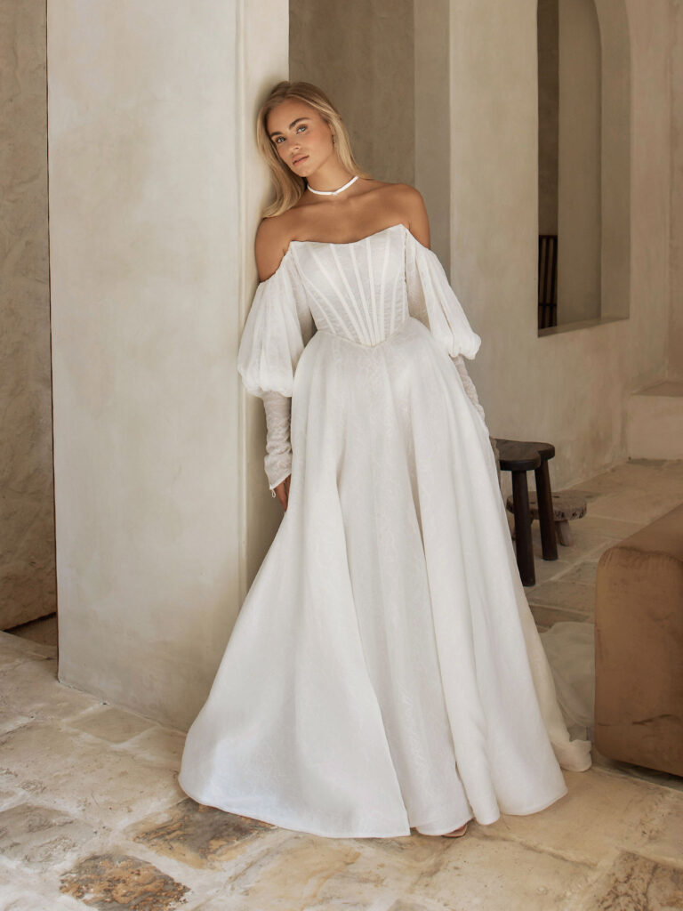 Sam-Serene by Madi Lane-Pure Bridal-Designed with vintage-inspiration for our more traditional brides, in modern fabrics to seamlessly blend the classic and contemporary. Our Sam wedding dress is both romantic and dramatic, ideal for making an unforgettable bridal statement.  Crafted in chiffon layered with Chantilly lace to create a soft, textural look.  A peaked, scoop neckline begins the bodice with a modern shape, the neckline continuing into the voluminous, off-shoulder blouson sleeves. These unique sleeves are fitted through the forearm in a cuff, coming to a point and covering the hand.  The bodice is designed sheer and illusionary, with contemporary exposed boning. The lines of which draw in the figure with the Basque waistline for a flattering shape.  A flat-front A-line skirt is given soft volume around the hem, keeping the look airy yet decadent. Extending around the back into the full, perfectly bridal train completed with a vertical row of buttons. Gracefully following in the wake of the bride.  The look is finished with a corset-style lace up back that feels classical and sensual. While also making it possible to further cinch in the waistline.  Style this dress with the delicate beaded Sam collar necklace. A sweet and timeless accessory to complete any wedding day look.